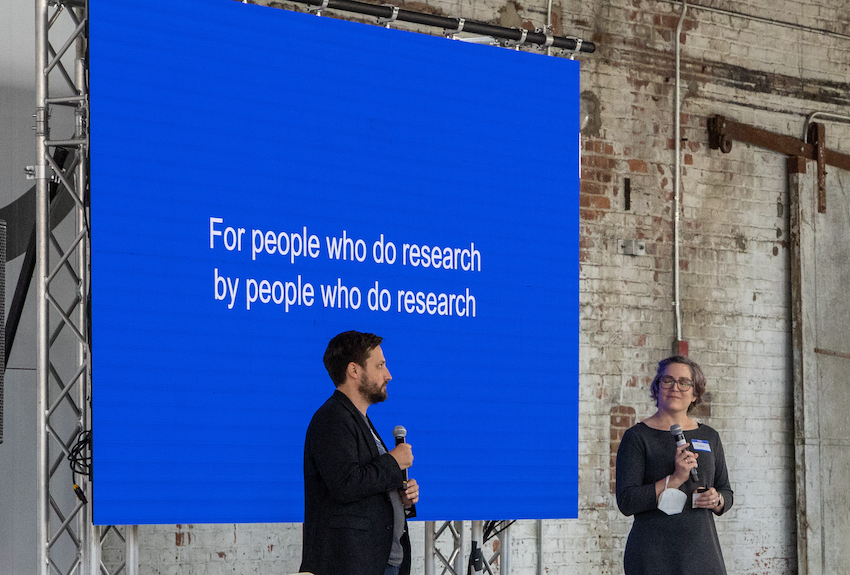 A man and a woman stand in front of a large screen displaying the words For people who do research, by people who do research on it