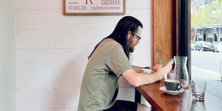 A man with long dark hair sitting alone in a coffee shop diligently working on his computer